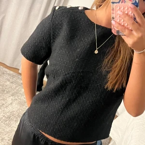 Svart kortärmad topp med knappar - Snygg svart topp med korta ärmar och dekorativa knappar på axlarna. Tillverkad i ett texturerat material som ger en elegant look. Storlek L men passar även M/S