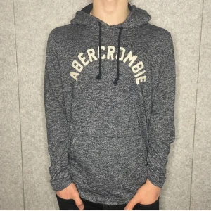 Grå hoodie från Abercrombie & Fitch - Säljer en snygg gråmelerad hoodie från Abercrombie & Fitch med deras logga i vitt på bröstet. Tröjan har en justerbar huva med snören och långa ärmar. Perfekt för en avslappnad stil. Nyskick 10/10 ny pris ca 1200kr 