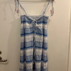 Blåvit klänning från Björn Borg - Säljer en somrig blåvit klänning från Björn Borg med smala axelband och knytning framtill. Klänningen har ett randigt mönster med dekorativa detaljer och är perfekt för varma dagar. 