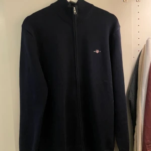 Mörkblå tröja med dragkedja från GANT - Snygg mörkblå tröja från GANT med dragkedja och ribbad krage. Perfekt för kyliga dagar. Den har en diskret logga på bröstet och är tillverkad i ett mjukt material. Tröjan är helt ny, prislapp finns kvar! Storlek M. Nypris 1699kr
