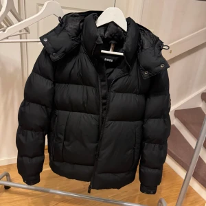 Svart pufferjacka från Boss - Snygg svart pufferjacka från Boss med dragkedja och avtagbar huva. Huvan har ett snyggt mönster från boss. Jackan har en quiltad design och en diskret logotyp på ärmen. Perfekt för kyliga dagar. Knappt använd! Storlek: 46 motsvarar S/M. Nypris 5000
