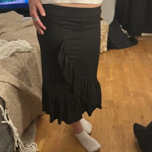 Svart kjol med volangdetaljer - Snygg svart kjol med volangdetaljer längs sidan och nederkanten. Kjolen har en omlott-design som ger en elegant touch. Perfekt för att klä upp eller ner beroende på tillfälle. ( instabox endast)