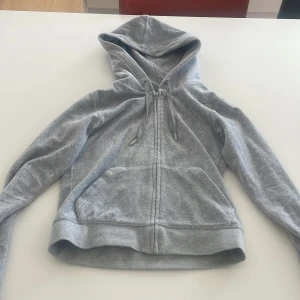 Grå hoodie från Juicy Couture - Säljer en stilren grå hoodie från Juicy Couture med dragkedja och praktiska fickor framtill. Perfekt för en avslappnad look. Huvan har justerbara snören med snygga metalldetaljer.