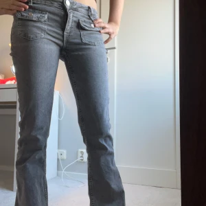 Grå jeansbyxor med fickdetaljer - Snygga grå jeansbyxor med låg midja och raka ben. Byxorna har unika fickdetaljer med knappar framtill och en klassisk dragkedja. Perfekta för en avslappnad stil.