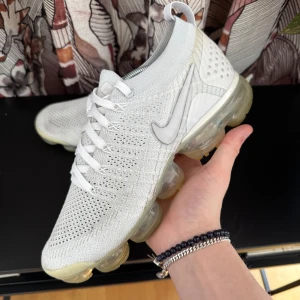 Nike Vapormax  - Snygga vita Nike Vapormax sneakers med stickad ovandel och transparent sula. Skorna har en modern design med snörning och en luftig känsla tack vare det stickade materialet. Perfekta för en sportig look. Spricka i sulan på ena skon.