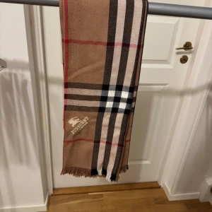 Burberry - Snygg rutig halsduk från Burberry i klassiska färger. Den har ett stilrent mönster med inslag av beige, svart, vit och röd. Perfekt accessoar för att lyfta din outfit. Har fått den av en vän så har inget kvitto eller liknande. Misstänker att den är oäkta.