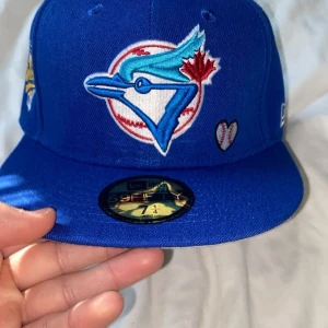 Blå keps från New Era - Snygg blå keps från New Era med broderad logotyp på framsidan och texten 'Love & Hustle = Game' på sidan. Kepsen har en platt skärm och är en del av 59FIFTY-serien. Perfekt för sportiga tillfällen.