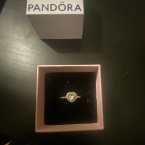 Pandora guld ring med hjärta - Superfin ring från Pandora i guldton med ett glittrande hjärta i mitten, omgiven av små stenar som ger extra shine. Klippt i bak av mig pga för stor storlek, man kan ändra det såklart. Fick av min gammla pojkvän använd runt 5 gånger🤍