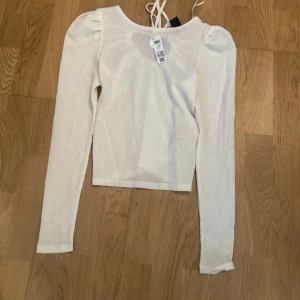Vit långärmad topp med knyt i ryggen - Superfin vit långärmad topp från Lindex i crinklat material. Toppen har puffiga axlar och snygg knytning i ryggen som ger en öppen rygg-effekt. Perfekt att styla med jeans eller kjol för en trendig look. Ny med prislapp