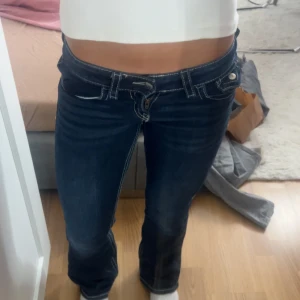 Mörkblå bootcut jeans med vita sömmar - Säljer ett par mörkblå jeans med bootcut passform och låg midja. Jeansen har tydliga vita kontrastsömmar och snygga fickdetaljer bak med knapp. Perfekta för dig som gillar klassisk denim med lite edge.