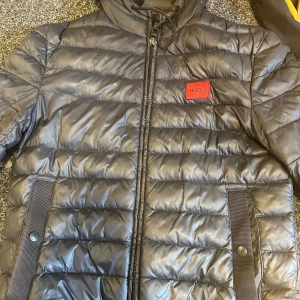 Svart pufferjacka från Hugo - Snygg svart pufferjacka från Hugo med quiltad design och röd logotyp på bröstet. Jackan har dragkedja framtill, två fickor med knappdetaljer och hög krage. Perfekt för kalla dagar och ger en clean streetstyle-vibe.