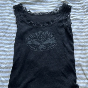 Svart ribbad linnetopp med spets - Svart ribbad linnetopp med spetskant runt halsringningen och ärmhålen. Storlek står inte men skulle gissa på Xs/S. Tryck framtill med text. Toppens material känns stretchigt och bra🫶🏻ALDRIG ANVÄND