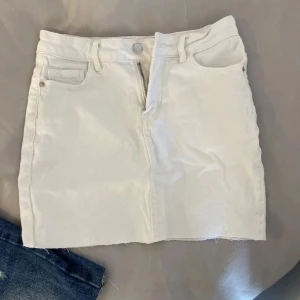 Vit jeanskjol med rå kant - Snygg vit jeanskjol med klassiska fickor fram och bak, bälteshällor och rå, avklippt kant nertill för en trendig look. Kjolen har dragkedja och knapp framtill. Perfekt för dig som gillar en clean och stilren vibe.