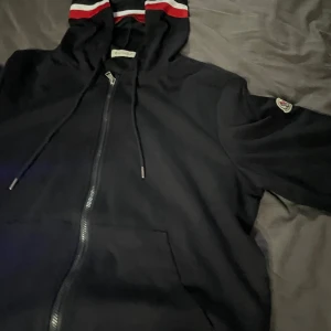 Svart Moncler hoodie - Svart hoodie från Moncler med hel dragkedja, huva med röd och vit randig detalj, samt Moncler-logga på ärmen. Tröjan har snörning i huvan och är långärmad. Perfekt för dig som gillar exklusiva streetwear-vibes och snygga detaljer.