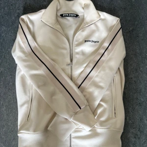 Beige/vit Palm Angels - Snygg beige track jacket från Palm Angels. dragkedja framtill, och två fickor med dragkedja. beige och svarta ränder längs ärmarna. Beige/vit resår i ärmar och midjan så den sitter åt o blir inte hängandes. 
