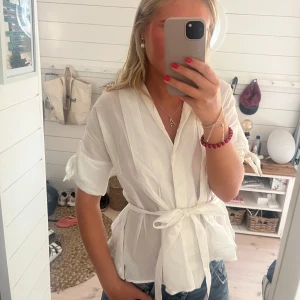 Vit Zara topp - Snygg vit omlottblus med knytband i midjan och korta ärmar med knyt-detalj. Luftigt och lätt material, perfekt för varma dagar. Stilren v-ringning och avslappnad passform som ger en fräsch och trendig look. Bomull