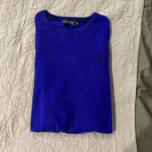 Ralph lauren stickad tröja - Snygg blå stickad tröja från Polo Golf Ralph Lauren i storlek M. Tröjan har rund hals, långa ärmar och en diskret svart logga broderad på bröstet. Perfekt för dig som gillar klassisk stil med en modern touch. Den är lite lila blå aktig en riktigt snygg och lite annorlunda färg som får dig att sticka ut från andra!