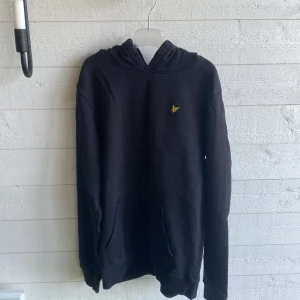Svart hoodie från Lyle & Scott - Svart hoodie från Lyle & Scott med klassisk gul fågel-logga på bröstet. Tröjan har huva, känguruficka och långa ärmar. Mjuk bomullskänsla och relaxed passform, perfekt för chill dagar. Enkel och stilren design som funkar till det mesta.