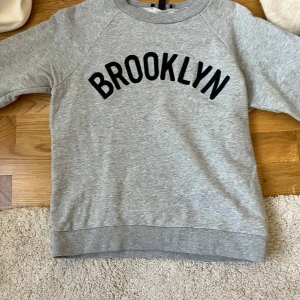 Grå sweatshirt Brooklyn från J.Crew - Säljer en grå sweatshirt från J.Crew i storlek S med svart BROOKLYN-tryck på bröstet. Tröjan har rund hals, långa ärmar och ribbade muddar vid hals, ärmslut och nederkant. Perfekt för chill dagar och streetstyle.