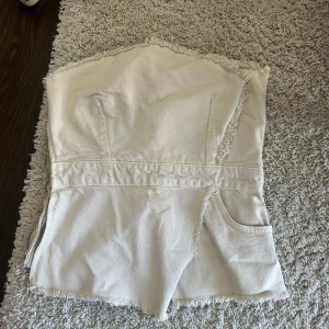 Beige jeans tubtopp med råa kanter - Säljer en beige tubtopp i jeanstyg med snygga råa kanter och en liten ficka framtill. Toppen är axelbandslös och har en cool, avslappnad vibe. Perfekt för dig som gillar en edgy och trendig stil.