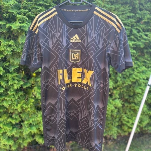 LAFC Adidas svart/guld matchtröja S - Snygg LAFC fotbollströja från Adidas i svart med guldiga detaljer och geometriskt mönster. Kortärmad modell med tre guldiga ränder på axlarna, klubbmärke och sponsortryck i guld framtill. Tillverkad i lätt och ventilerande polyester. Har ni några frågor om lappar eller annat är det bara att höra av sig! Priset är ej hugget i sten👍