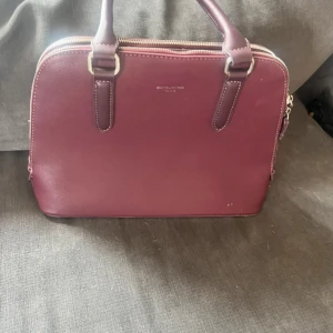 Vinröd handväska från David Jones - Snygg vinröd handväska från David Jones med stilren design och två rejäla handtag. Väskan har silverfärgade dragkedjor, ett ytterfack med dragkedja och rymligt beige innerfoder. Tillverkad i syntetmaterial och perfekt för dig som gillar klassisk look.