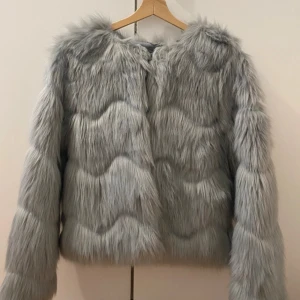 Ljusgrå pälsjacka med vågig struktur - Supermjuk och fluffig ljusgrå pälsjacka med vågig struktur i pälsen. Jackan har lång ärm, rund halsringning och stängs med hyska. Fodrad insida för extra komfort. Perfekt för dig som vill sticka ut med en trendig look. Använd bara en gång! 🎀