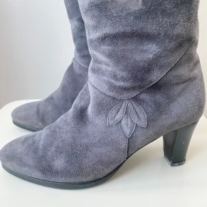 Grå mockastövlar med klack - Snygga grå stövlar i mocka från  juhani palmroth med medelhög klack och dekorativt bladmönster på sidan. Stövlarna har rund tå, dragkedja på insidan och en klassisk siluett som passar till många outfits.