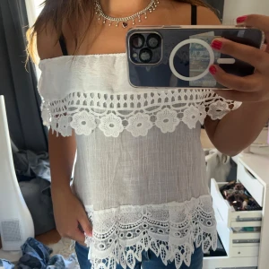 Vit offshoulder blus med spetsdetaljer - Superfin vit offshoulder blus med breda spetskanter och broderade blommor. Luftigt tyg och snyggt fall, perfekt till jeans eller kjol. Blusen har ärmlös design och vackra spetsmönster både upptill och nertill för en romantisk vibe.