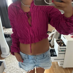 Fuchsia croppad blus med knyt - Säljer en fuchsiafärgad croppad blus med långa puffiga ärmar och snygga rynkade detaljer. Toppen har knyt framtill och volangkant nedtill för en trendig look. Perfekt för dig som vill sticka ut med färg och stil.
