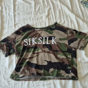 Camouflage t-shirt från SikSilk - Cool croppad t-shirt från SikSilk med tydligt broderad logga på bröstet. Camouflage-mönster i grönt, brunt, beige och svart. Rund hals och korta ärmar. Perfekt för dig som gillar streetwear och vill sticka ut med en edgy stil.