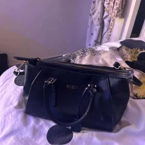 Svart handväska från Guess - Snygg svart handväska från Guess med guldfärgade detaljer och dragkedja upptill. Väskan har både handtag och en justerbar axelrem. Insidan är fodrad med beige tyg och Guess-logga. Perfekt storlek för att få plats med allt du behöver. Pris kan diskuteras ❤️