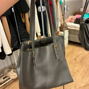 Mörkgrå axelväska i skinn - Snygg mörkgrå axelväska i skinn med justerbara axelband och guldfärgade spännen. Väskan har flera fack, inklusive ett med dragkedja, vilket gör den perfekt för att hålla ordning på dina grejer. Stilren och rymlig design som passar till det mesta.