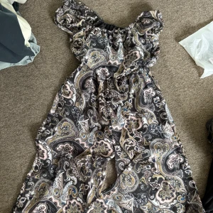 Svart paisleymönstrad jumpsuit  - Säljer en svart jumpsuit med paisleymönster i beige, blått och ljusrosa. Jumpsuiten har offshoulder-ärmar med resår och ett luftigt fall. Materialet känns som polyester och har en mjuk innersida. Perfekt för dig som gillar boho-vibe och detaljerade mönster.