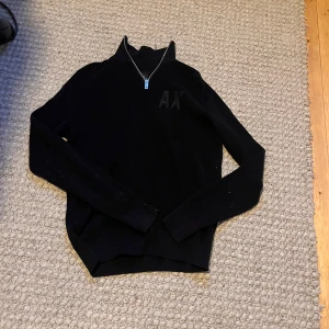 Svart stickad tröja Armani Exchange - Svart stickad tröja från Armani Exchange med half zip och högt krage. Diskret AX-logga på bröstet och ribbade muddar vid ärmslut och nederkant. Perfekt för dig som gillar stilrena och klassiska plagg med en modern touch. Bara att höra av sig med frågor!