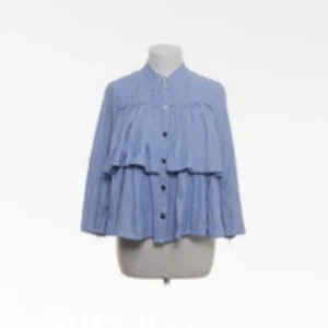 Blå volangskjorta med knappar - Supersöt blå skjorta med volanger och knäppning framtill. Skjortan har en lös passform och lång ärm, perfekt för dig som gillar en chill och trendig stil. Ståkrage och lätta material gör den extra bekväm.