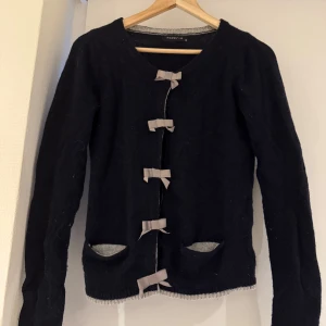 Mörkblå kofta med rosetter från Modström - Supersöt svart kofta från Modström med grå detaljer och fyra grå rosetter framtill. V-ringad och med långa ärmar, samt två fickor med grå insida. Mjuk stickad känsla och knäppning med både rosetter och dolda tryckknappar. Perfekt för dig som gillar gulliga detaljer!