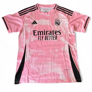 Rosa Real Madrid fotbollströja Adidas - Snygg rosa Real Madrid fotbollströja från Adidas med svart logga och svarta detaljer på axlar och sidor. Tröjan har korta ärmar, rund hals och tryck med Emirates Fly Better på bröstet. Tillverkad i lätt och ventilerande material, perfekt för träning eller match.