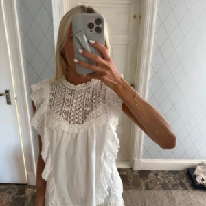 Vit blus med spets och volang - Superfin vit blus med spetsdetaljer upptill och volanger längs axlarna. Hög halsringning och ärmlös design ger en romantisk vibe. Perfekt till jeans eller kjol för en söt look. Luftigt bomullsmaterial som känns fräscht hela dagen.