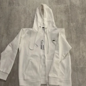 Lacost tracksuit - Säljer en fräsch vit hoodie från Lacoste med klassisk krokodillogga på bröstet. Tröjan har huva med snörning, praktiska fickor och dragkedja framtill. Perfekt för chill dagar och snygg att matcha med jeans eller joggers. Materialet är mjuk bomull som känns skönt mot huden.