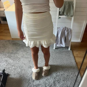 Vit kortkjol med volangkant - Supersöt vit kortkjol med volangkant längst ner. Kjolen har en tight passform och är gjord i mjukt bomullsmaterial som sitter skönt mot kroppen. Perfekt för dig som gillar en feminin och fräsch stil. Enkel att matcha med crop tops eller stickade tröjor.