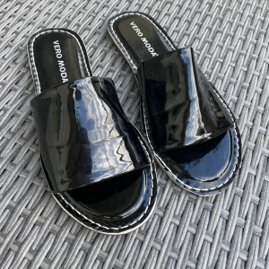 Svarta sandaler från Vero Moda - Svarta sandaler från Vero Moda med glansig finish och bred rem över foten. Platt sula med vita kontrastsömmar runt kanten. Öppen tå och enkel slip-in modell, perfekta för varma dagar.Slip on Sandaler i lack från veromoda. Använda 1-2 gånger men mycket Fint skick. Storlek 37, normal i storlek. Jag har massor av artiklar till salu! Köp flera och få rabatt!