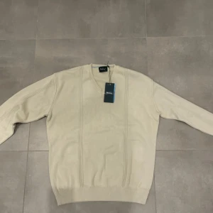 Beige v-ringad tröja från Wolsey - Snygg beige v-ringad tröja från Wolsey med diskreta ribbade detaljer längs fram och på ärmarna. Tröjan är långärmad och har en klassisk, normal passform. Perfekt för dig som gillar stilrena och enkla plagg med lite extra struktur. Materialet är mjukt och känns skönt mot huden.