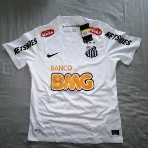 Santos Neymar Jr vit Nike fotbollströja - Snygg vit Santos FC matchtröja från Nike med Neymar Jr och nummer 11 på ryggen. Tröjan har korta ärmar, rund hals och flera tryckta sponsorer i svart och orange. Materialet är lätt och andas, Dri-Fit polyester. Perfekt för dig som älskar brasiliansk fotboll.