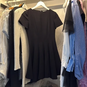 Svart kortklänning från Zara XS - Snygg svart klänning från Zara i storlek XS. Modellen är kort med rund hals och korta ärmar, har en lätt utställd kjol och är tillverkad i ett mjukt syntetmaterial. Perfekt för dig som gillar stilrena och enkla plagg med modern passform.