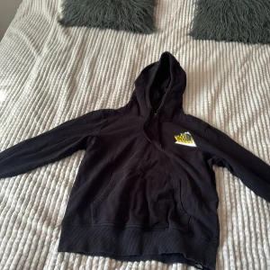 Svart hoodie från The North Face - Svart hoodie från The North Face med klassisk logga i gult och vitt på bröstet. Tröjan har huva med snörning, stor magficka och ribbade muddar vid ärmslut och nederkant. Perfekt för chill dagar eller när du vill ha en skön streetstyle.