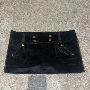 Svart manchesterkortkjol med guldknappar - Supersnygg svart kortkjol i mjuk manchester med guldiga knappar framtill och på fickorna. Kjolen har praktiska fickor både fram och bak samt dragkedjor i sidorna. Perfekt för dig som gillar detaljer och vill ha en cool look. Shorts under❤️