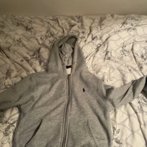 Grå hoodie från Polo Ralph Lauren - Säljer en grå hoodie från Polo Ralph Lauren med dragkedja och huva. Tröjan har ett litet lila Polo-logo broderat på bröstet, fickor fram och mjukt vitt fleecefoder på insidan. Perfekt för chill dagar och snygg till jeans.