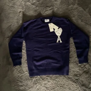 Ami Paris tröja - Snygg mörkblå stickad tröja från Ami Alexandre Mattiussi med stor vit hjärtlogga och bokstaven A på bröstet. Klassisk rund halsringning och långa ärmar. Perfekt för dig som gillar stilrena plagg med en twist.