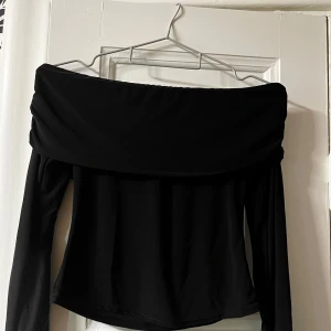 Svart offshoulder topp från Boohoo(I liknande satin material) - Säljer en svart offshoulder topp från Boohoo i stretchigt material. Toppen har långa ärmar och en bred, vikt kant över axlarna som ger en snygg och trendig look. Perfekt att styla med jeans eller kjol för en cool outfit.